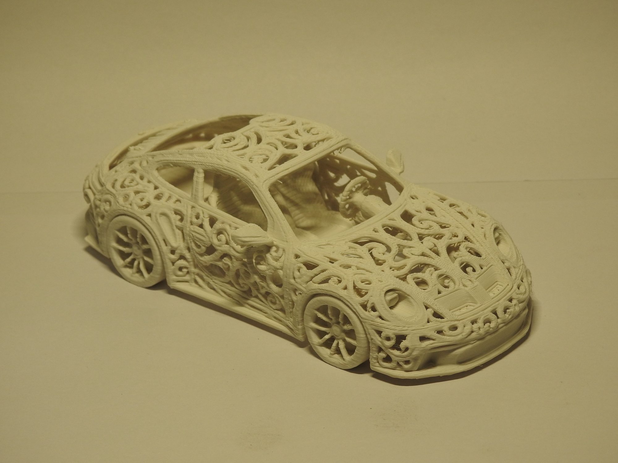 Ivory 3D printed Porsche 992 GT3 seen from above the front left corner, with a swirling Voronoi lattice across the hood and roof and the seats and steering wheel visible inside